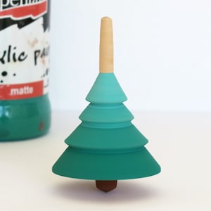 Puede incluir: Una peonza de madera con forma de árbol de Navidad, pintada en tonos verde y verde azulado. La parte superior y la base son de madera natural. En el fondo se ve un bote de pintura acrílica verde mate.