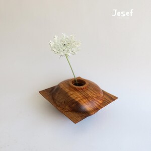 May include: A handcrafted wooden vase with a square base and a rounded top, holding a single white flower. The vase is made of a light brown wood with visible grain patterns. The Josef Wood Works logo is in the top right corner.
