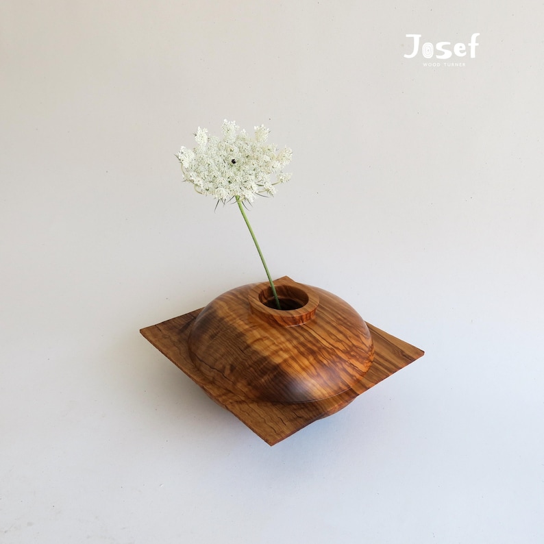 May include: A handcrafted wooden vase with a square base and a rounded top, holding a single white flower. The vase is made of a light brown wood with visible grain patterns. The Josef Wood Works logo is in the top right corner.
