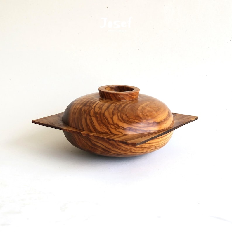 May include: A handcrafted wooden bowl with a lid. The bowl is made of olive wood, featuring a natural wood grain pattern. The lid has a small, cylindrical handle. The bowl sits on a square base, creating a unique design.
