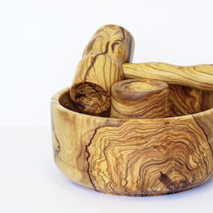 May include: A wooden mortar and pestle set with a bowl, a pestle, and a small cylindrical holder. The set is made of olive wood, showcasing a natural grain pattern in shades of brown and tan. The bowl is round and the pestle has a rounded head.