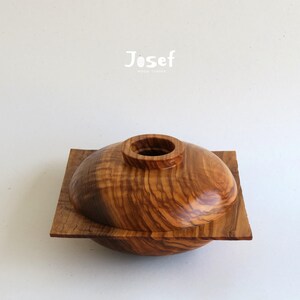May include: A handcrafted wooden bowl with a lid, featuring a natural wood grain pattern. The bowl is set on a square base, and the lid has a cylindrical opening. The word "Josef" is printed on the top.