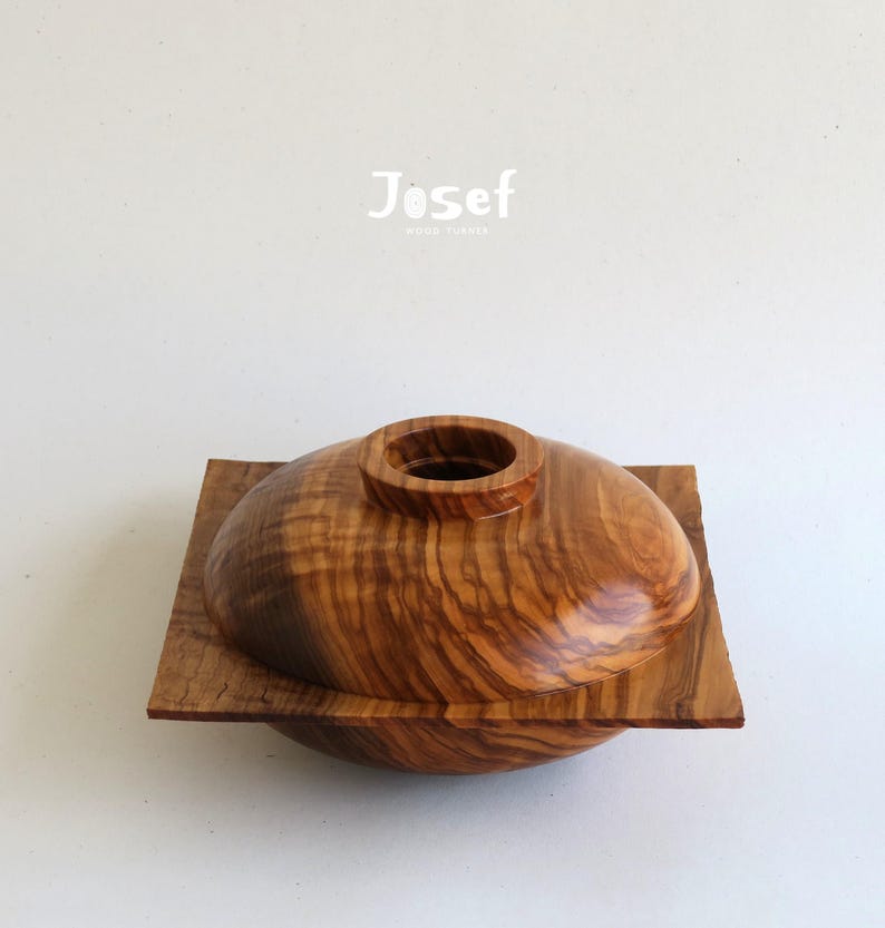 May include: A handcrafted wooden bowl with a lid, featuring a natural wood grain pattern. The bowl is set on a square base, and the lid has a cylindrical opening. The word "Josef" is printed on the top.