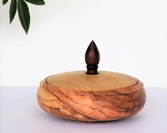 May include: A wooden bowl with a lid and a dark wood knob on top. The bowl is made of olive wood and has a natural wood grain pattern.