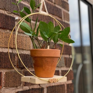Peut inclure: Un pot de fleurs suspendu en métal doré avec une étagère en bois et les lettres "P C O" dessus. Une plante verte dans un pot en terre cuite est suspendue dans le pot de fleurs.