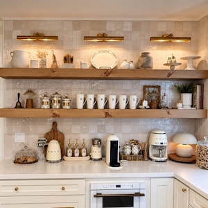 May include: A kitchen featuring white cabinets with gold hardware, two natural wood floating shelves, and a white countertop. The shelves hold various ceramic mugs, jars, and decorative items. A white microwave, coffee maker, and kettle are present.