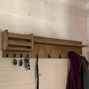 May include: A wooden wall-mounted coat rack with a shelf and a small cubby. The rack has six hooks and is stained a dark brown color. There are keys and a leash hanging from the hooks, and a purple jacket and a black jacket are hanging on the hooks.