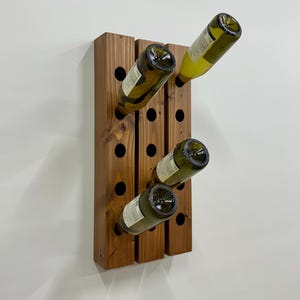 May include: A wall-mounted wine rack constructed from three vertical dark brown wooden beams with circular cutouts. Three glass wine bottles are inserted into the holes at an upward angle, showcasing the natural wood grain and texture.