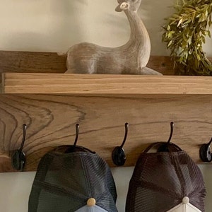 May include: A wooden shelf with five black metal hooks, two baseball caps hanging on the hooks, and a wooden deer figurine on the shelf.