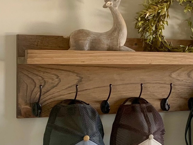 May include: A wooden shelf with five black metal hooks, two baseball caps hanging on the hooks, and a wooden deer figurine on the shelf.