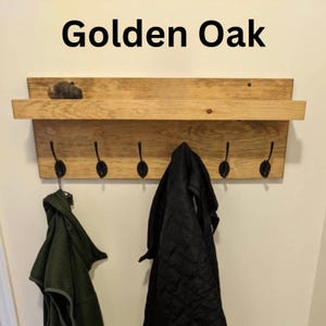 以下が含まれることがあります： Golden Oakという文字が記された、棚付きの木製壁掛けコートラック。木目がはっきりと見える明るい茶色の木材で作られており、5つの黒い金属製フックが付いています。オリーブグリーンの衣類と黒いキルティングジャケットがフックに掛けられています。