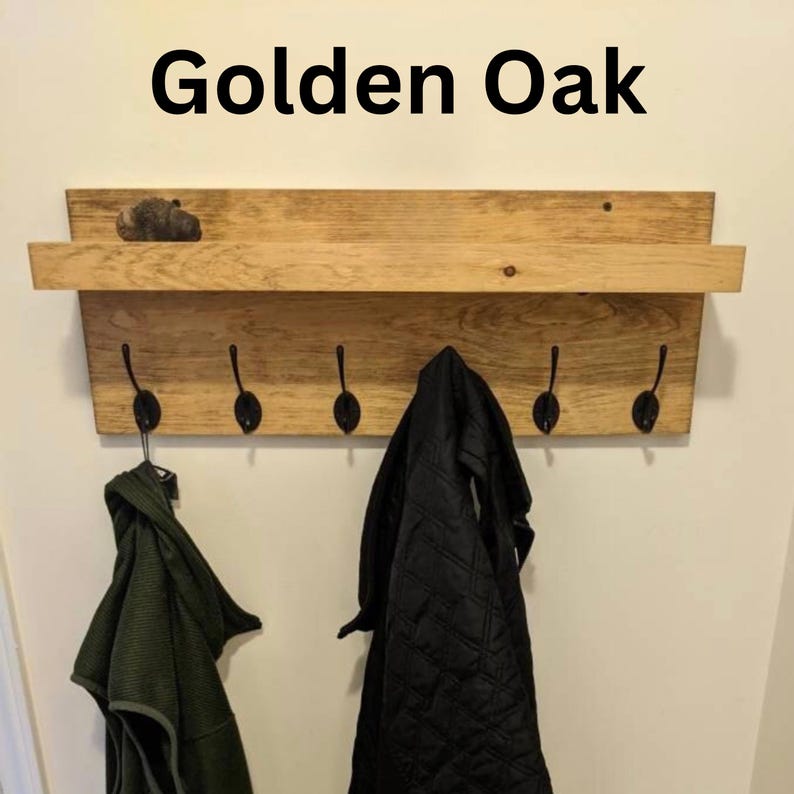 以下が含まれることがあります： Golden Oakという文字が記された、棚付きの木製壁掛けコートラック。木目がはっきりと見える明るい茶色の木材で作られており、5つの黒い金属製フックが付いています。オリーブグリーンの衣類と黒いキルティングジャケットがフックに掛けられています。
