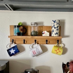 May include: A wooden shelf with five hooks, holding four mugs and a glass jar. The mugs have various designs, including a blue and white star, a yellow mug with a red sun symbol, and a white mug with the text "This I Like The Most Call Me Papa Frank". The jar is clear glass with a metal lid and contains dried herbs.