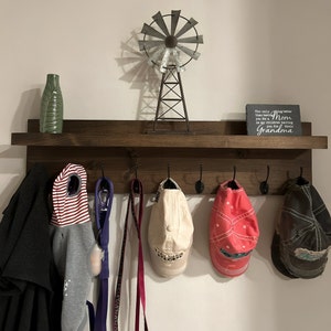 May include: A dark brown wooden shelf with six hooks and a small metal windmill on top. The shelf is mounted on a white wall. There are various items hanging on the hooks, including a red and white striped hooded sweatshirt, a purple leash, a pink leash, a beige baseball cap, a red baseball cap, and a brown baseball cap. A small gray sign with white text reads "The only thing better than being your Mom is my children having you for their Grandma".