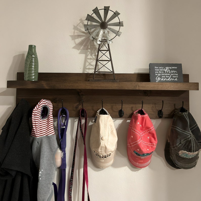 May include: A dark brown wooden shelf with six hooks and a small metal windmill on top. The shelf is mounted on a white wall. There are various items hanging on the hooks, including a red and white striped hooded sweatshirt, a purple leash, a pink leash, a beige baseball cap, a red baseball cap, and a brown baseball cap. A small gray sign with white text reads "The only thing better than being your Mom is my children having you for their Grandma".
