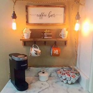 May include: A rustic coffee bar setup with a wooden shelf, hooks, and a sign that reads "coffee bar". There is a Keurig coffee maker, a white ceramic sugar bowl, and a metal basket filled with coffee pods.