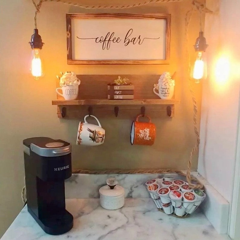 May include: A rustic coffee bar setup with a wooden shelf, hooks, and a sign that reads "coffee bar". There is a Keurig coffee maker, a white ceramic sugar bowl, and a metal basket filled with coffee pods.