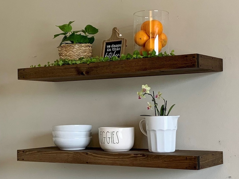 May include: Two dark brown floating shelves with decorative items. The top shelf has a potted plant, a small cutting board with text, and a glass vase with oranges. The bottom shelf displays white bowls and a ceramic bowl with the word "VEGGIES".