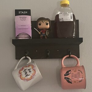 May include: A black shelf with three hooks holds a mug with the words "be kind" in rainbow colors, a pink mug with the words "protect your peace" in gold lettering, a box of Stash Breakfast in Paris black tea, and a bottle of raw local honey.