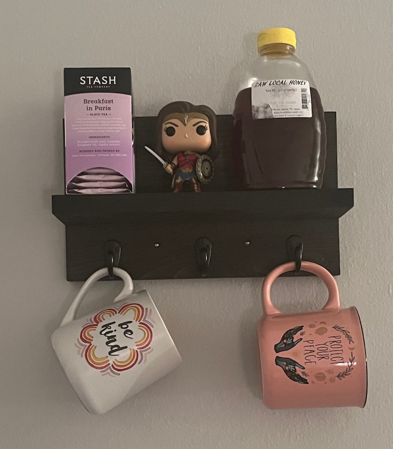 May include: A black shelf with three hooks holds a mug with the words "be kind" in rainbow colors, a pink mug with the words "protect your peace" in gold lettering, a box of Stash Breakfast in Paris black tea, and a bottle of raw local honey.