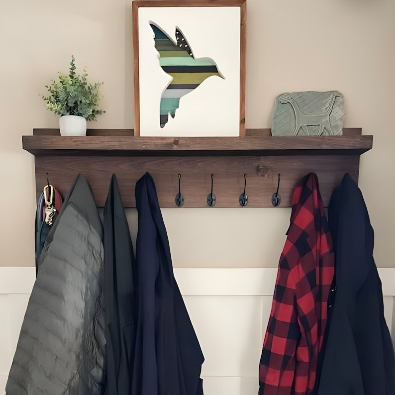 May include: A dark brown wooden coat rack with six hooks and a shelf. The shelf has a white framed picture with a green and blue hummingbird silhouette. There are five coats hanging on the hooks, including a red and black plaid coat.