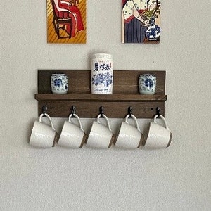 May include: A dark brown wooden shelf with five hooks and two small decorative ceramic jars. The shelf is mounted on a white wall. Five white ceramic mugs hang from the hooks.