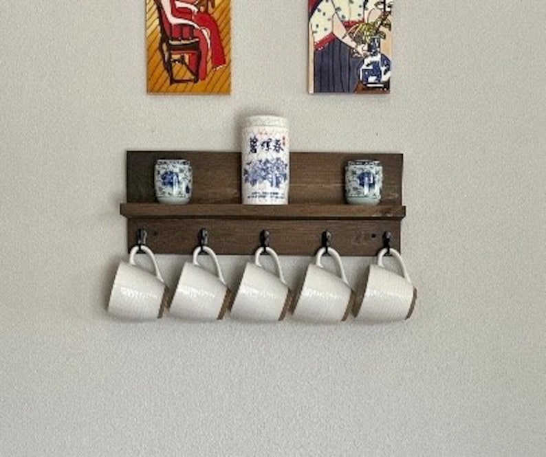 May include: A dark brown wooden shelf with five hooks and two small decorative ceramic jars. The shelf is mounted on a white wall. Five white ceramic mugs hang from the hooks.