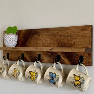 May include: A wooden shelf with hooks for hanging mugs. The shelf is stained a dark brown color and has a small plant in a white pot on the top shelf. There are six mugs hanging from the hooks, each with a different butterfly design.