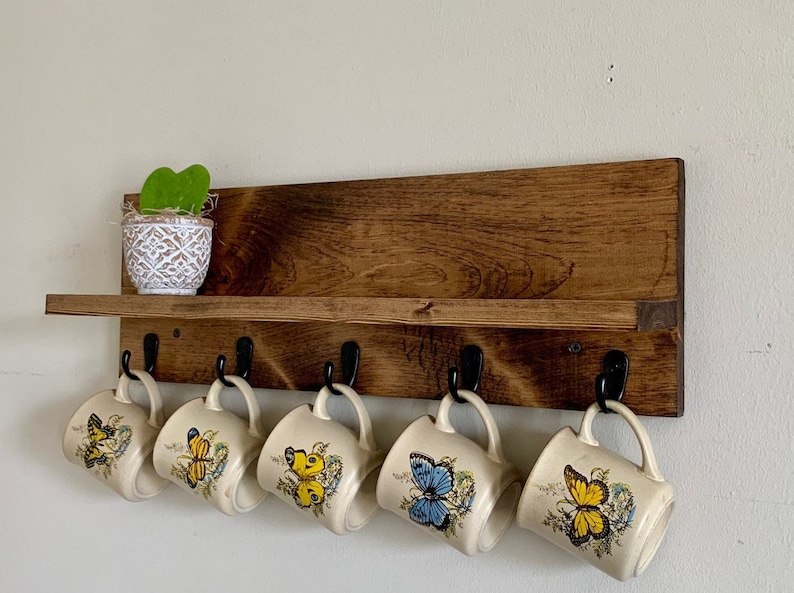 May include: A wooden shelf with hooks for hanging mugs. The shelf is stained a dark brown color and has a small plant in a white pot on the top shelf. There are six mugs hanging from the hooks, each with a different butterfly design.