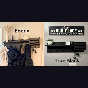 May include: Two wooden coat racks with shelves and hooks. The rack on the left is stained in a dark brown color labeled "Ebony". The rack on the right is painted black and labeled "True Black". Both racks have a sign above them that reads "Welcome to Our Place Friends & Family Gather Here".