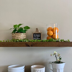May include: A wooden shelf features a small potted plant in a woven basket, a sign reading "We dance in this kitchen", and a glass jar filled with oranges. A green vine and a white pitcher with a flower arrangement complete the display. The shelf is approximately 120 cm long.