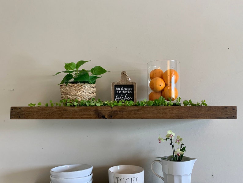 May include: A wooden shelf features a small potted plant in a woven basket, a sign reading "We dance in this kitchen", and a glass jar filled with oranges. A green vine and a white pitcher with a flower arrangement complete the display. The shelf is approximately 120 cm long.