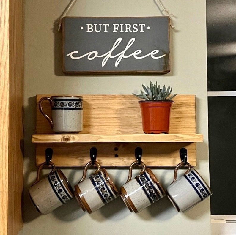May include: A wooden shelf with five hooks for hanging mugs. The shelf has a sign that reads "But First, Coffee". There are four mugs hanging on the hooks and one mug sitting on the shelf. The mugs are all white with blue and brown designs.