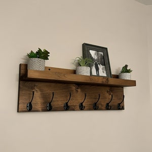 May include: A wooden shelf with six black metal hooks and a brown stained finish. The shelf has three potted plants and a framed photo on top.