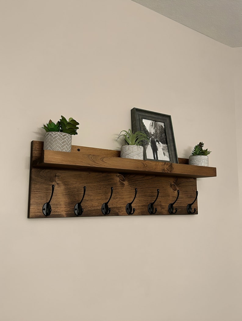 May include: A wooden shelf with six black metal hooks and a brown stained finish. The shelf has three potted plants and a framed photo on top.