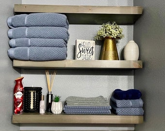 May include: Two wooden shelves with gray and blue towels, a sign that says "Hello Sweet Cheeks", a gold vase with green plants, a white vase, a red air freshener, a black candle holder, a bottle of lotion, and a small potted plant.