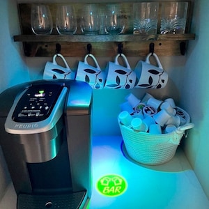 May include: A silver Keurig coffee maker with a digital display and a basket of coffee pods. The coffee maker is on a white countertop with a green and white sign that says "BAR" on it. There are also four white mugs with a leaf design hanging on a wooden shelf above the coffee maker.
