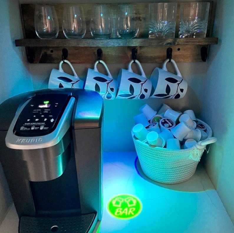 May include: A silver Keurig coffee maker with a digital display and a basket of coffee pods. The coffee maker is on a white countertop with a green and white sign that says "BAR" on it. There are also four white mugs with a leaf design hanging on a wooden shelf above the coffee maker.