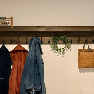 May include: A dark brown wooden coat rack with a shelf and multiple hooks. There are three jackets hanging on the hooks, a basket, a wreath, and a small wooden abacus on the shelf.