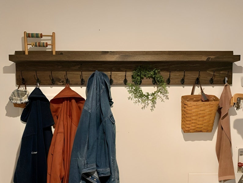 May include: A dark brown wooden coat rack with a shelf and multiple hooks. There are three jackets hanging on the hooks, a basket, a wreath, and a small wooden abacus on the shelf.