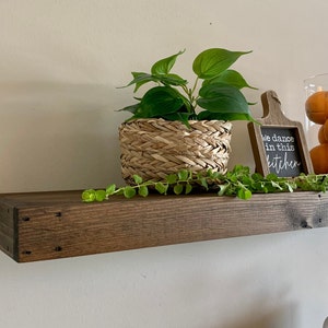 May include: A dark stained wooden shelf displays a woven basket with a green plant, a small chalkboard sign that reads "We dance in this kitchen", and a glass jar filled with oranges. A green vine trails across the shelf.