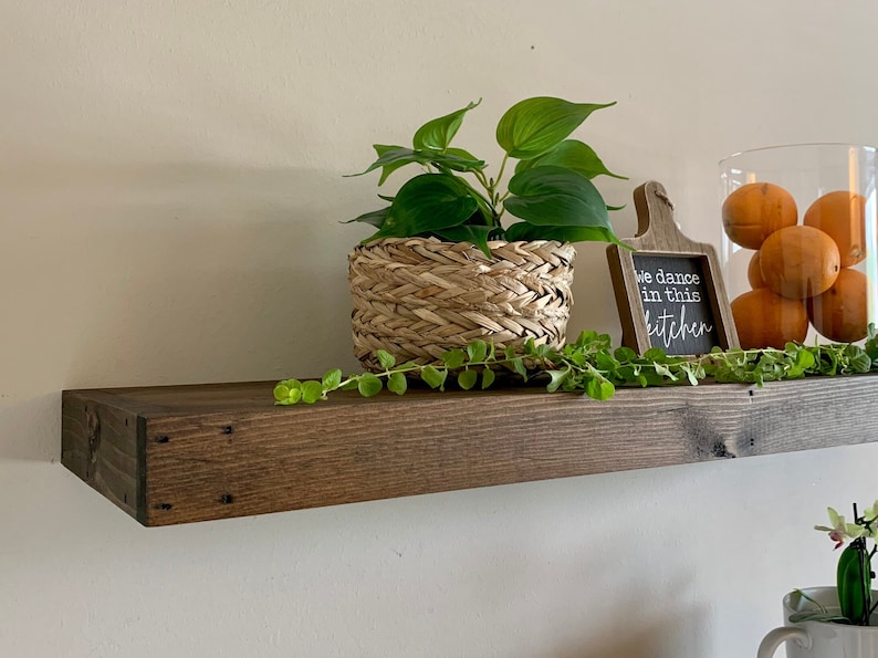 May include: A dark stained wooden shelf displays a woven basket with a green plant, a small chalkboard sign that reads "We dance in this kitchen", and a glass jar filled with oranges. A green vine trails across the shelf.