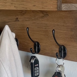 May include: A wooden wall-mounted shelf with three black metal hooks. Two jackets hang on the hooks, and keys are hanging from the bottom hooks.