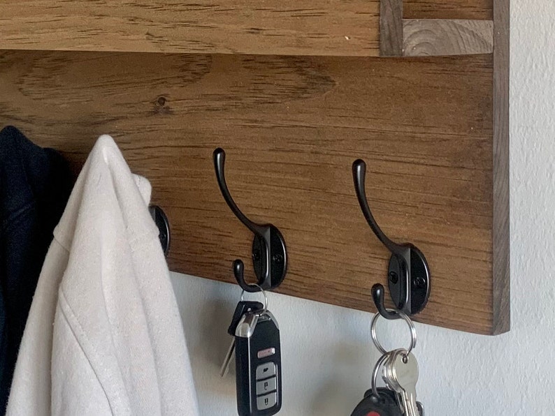 May include: A wooden wall-mounted shelf with three black metal hooks. Two jackets hang on the hooks, and keys are hanging from the bottom hooks.