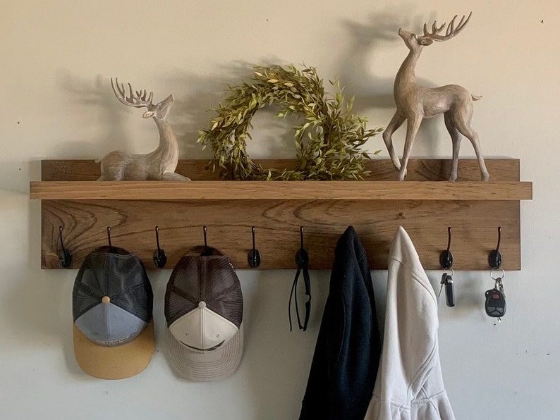 May include: A wooden shelf with five hooks, two hats, two coats, and two keychains hanging on it. There are two decorative deer figurines and a wreath on the shelf.