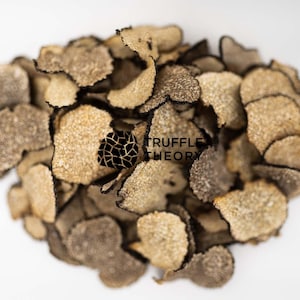 Puede incluir: Primer plano de un montón de rodajas de trufa negra seca. Las rodajas son de color marrón claro con bordes marrón oscuro. Las rodajas de trufa están dispuestas en un patrón aleatorio. El texto "TRUFFLE THEORY" es visible en la imagen.