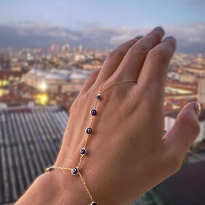 Op de afbeelding: Een gouden handketting met kleine blauwe en witte evil eye bedels. De fijne ketting verbindt met een armband en loopt door tot een ring. De sieraden worden getoond tegen een vage stadsachtergrond.
