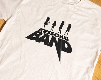 May include: A cream-colored short-sleeve t-shirt featuring a black graphic print on the chest. The design displays the text KESSOKU BAND in a bold, stylized font with four black silhouettes of figures standing above the lettering.