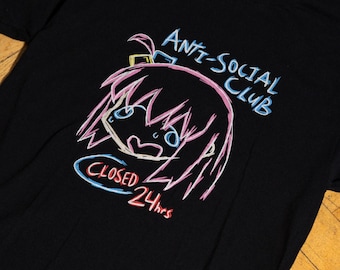 May include: A black t-shirt featuring a graphic print of a stylized anime-style character with pink hair. The text "ANTI-SOCIAL CLUB" appears in blue above the character, and the text "CLOSED 24hrs" is printed in red and blue below the character.
