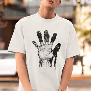 May include: White t-shirt with a black and white graphic of a hand with four fingers. Each finger has a small, grotesque face.